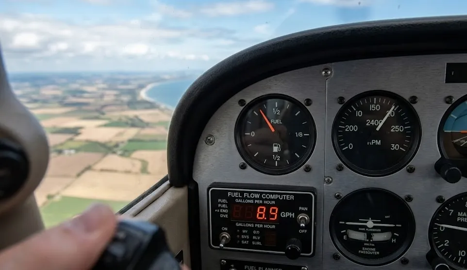 Cockpit view of a Cessna 172 dashboard showing a fuel flow computer and gauges over a landscape.